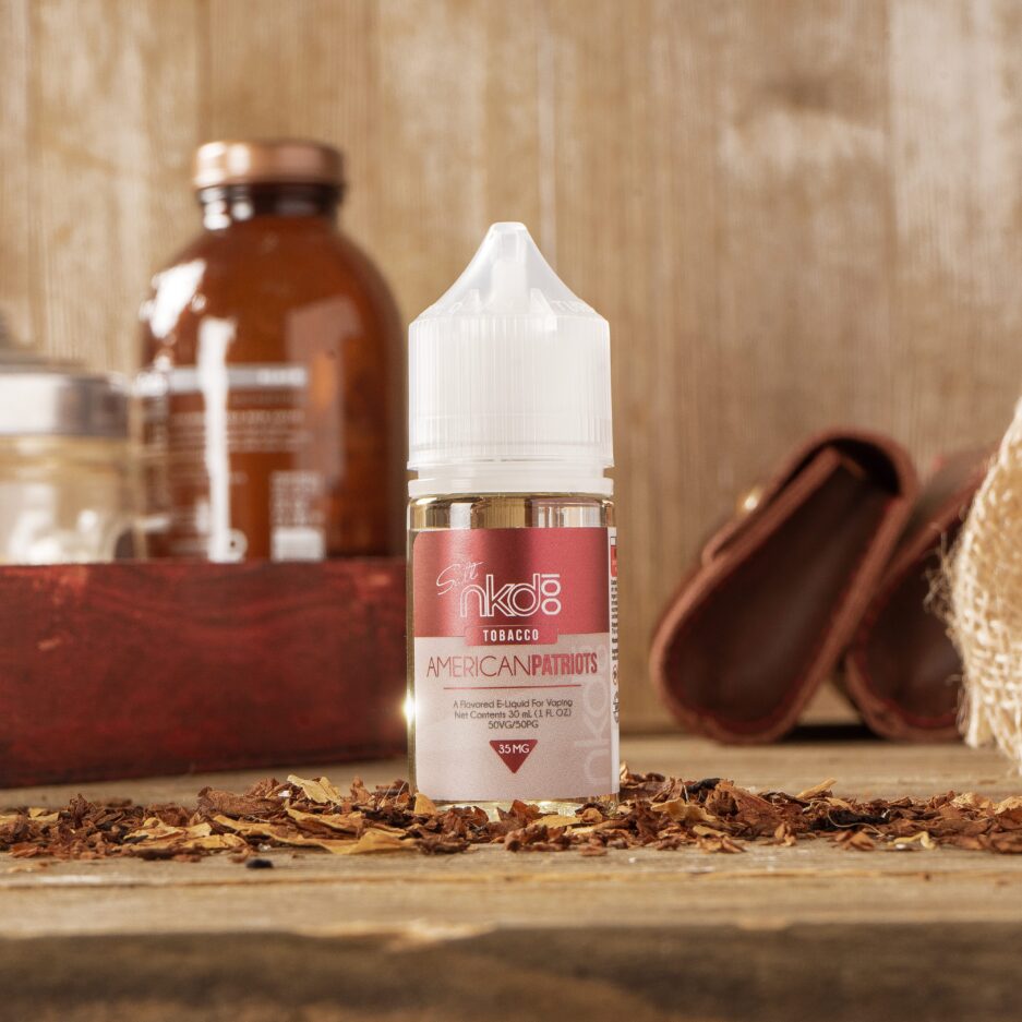 American Patriots standing at the center of a wooden table top setting with a wooden wall. Tobacco shavings are lined with the bottle horizontally partially covering the floor. In the background are random objects that give a sense of home and comfiness.