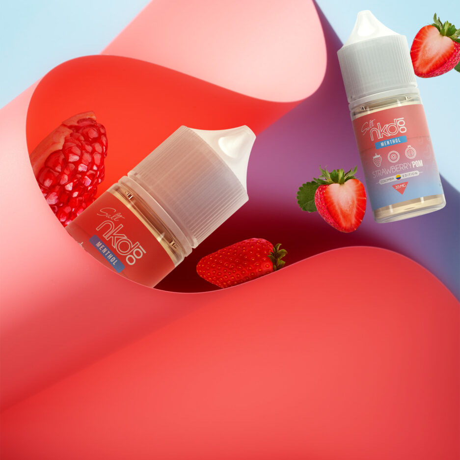 A red abstract object with a wavy paper like fold controls the image, partially in view is a Strawberry Pom bottle, and towards the right getting farther are strawberries and a pomegranate along with another bottle of Strawberry Pom flying as well