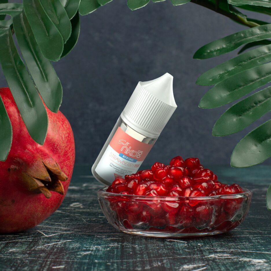 A dark colored wood textured floor and matte black wall, a leaf creating an arch on the top of the image, a glass bowl of pomegranate seeds and Strawberry Pom Salt angled next to the bowl.