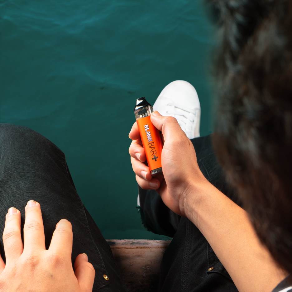 A top down angle of a man sitting down on a wooden dock with his feet hanging by the water holding a Blank Bar Plus Hybrid Disposable in his right hand.