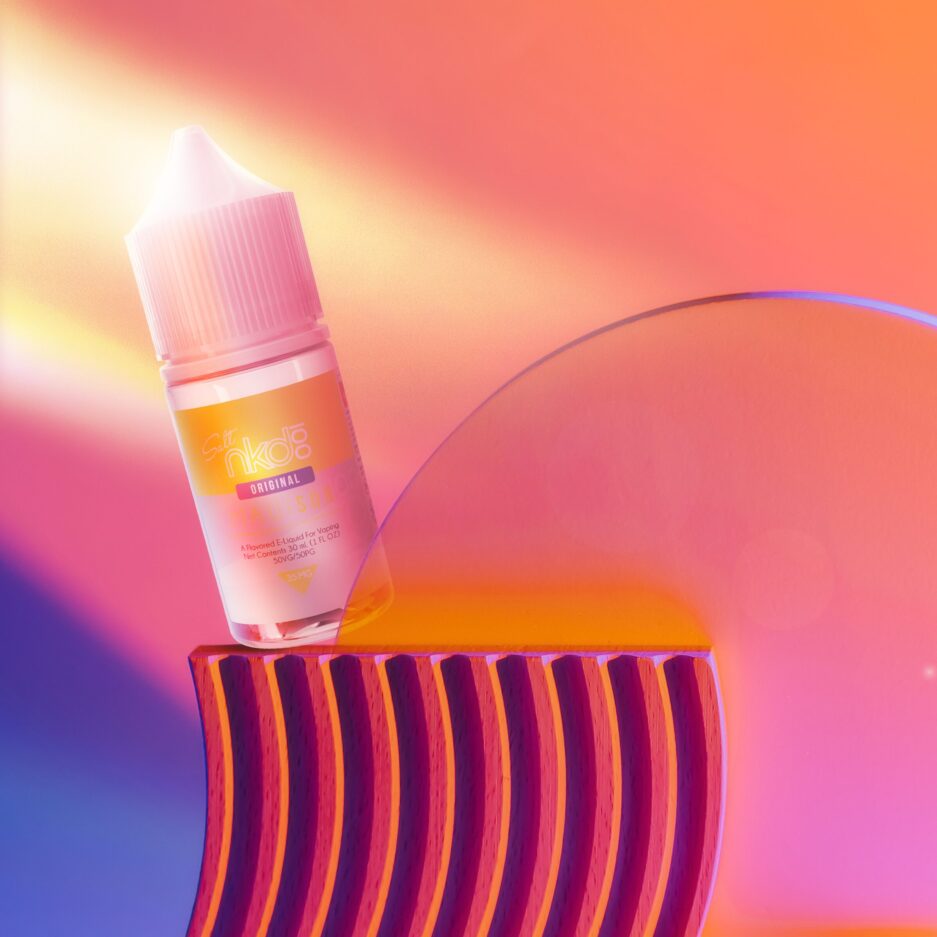 A product photo with a sunset color palate, light illuminating from the left side outside of the image creating a dramatic setup. two abstract objects sit front and center right of the image with Naked100 internationals Maui Sun sits on one of the objects, partially illuminated by the said light source.