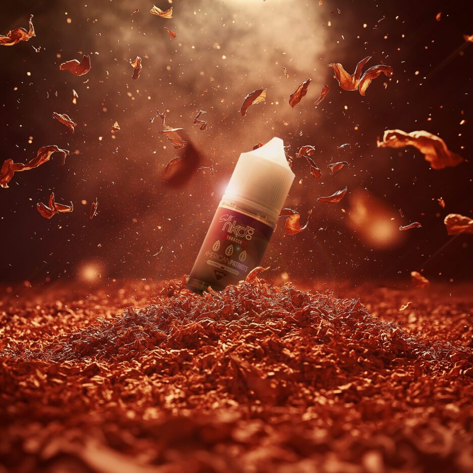 A product shot with red and burgundy color tones, a pile of tobacco covering the floor with one big pile at the center with tobacco leafs falling towards the center from the top, Naked100 Colombia's American Patriots sits in the center partially dug into the tobacco, with light from the top partially illuminating the bottle. Minimal smoke is rising in the air.