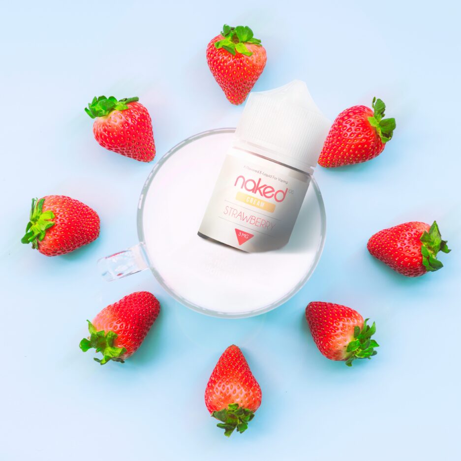 A top down product shot with a light baby blue floor, there is a glass in the center of the image filled to the brim with cream. Naked 100's Strawberry Cream 60mL bottle is sitting halfway inside the glass with the other half of the bottle peering out for the viewer to see. Strawberries are placed systematically around the glass and bottle, all pointing towards the center of the image.